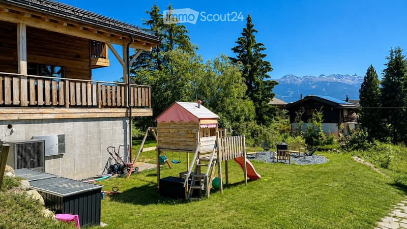 A well-maintained garden with a wooden playhouse, slide, swing, and benches, surrounded by trees and a view of mountains.