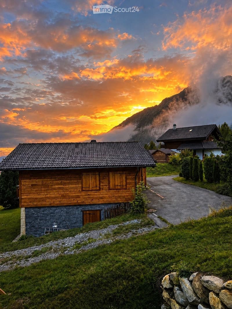 A wooden chalet with a stone base, situated on a grassy slope with a winding driveway leading up to it. The background features a mountain range and a dramatic sunset sky.