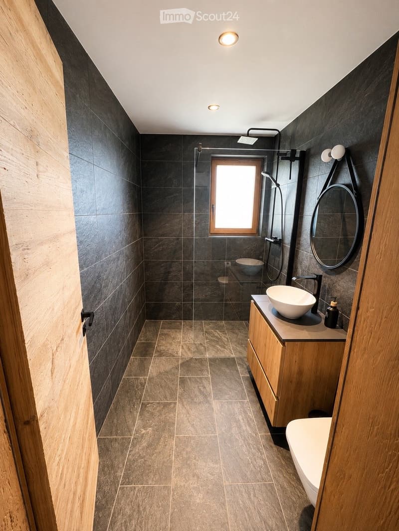 Modern bathroom with dark grey tiles, a large window, a wooden vanity with a sink, a bathtub, and a shower area.