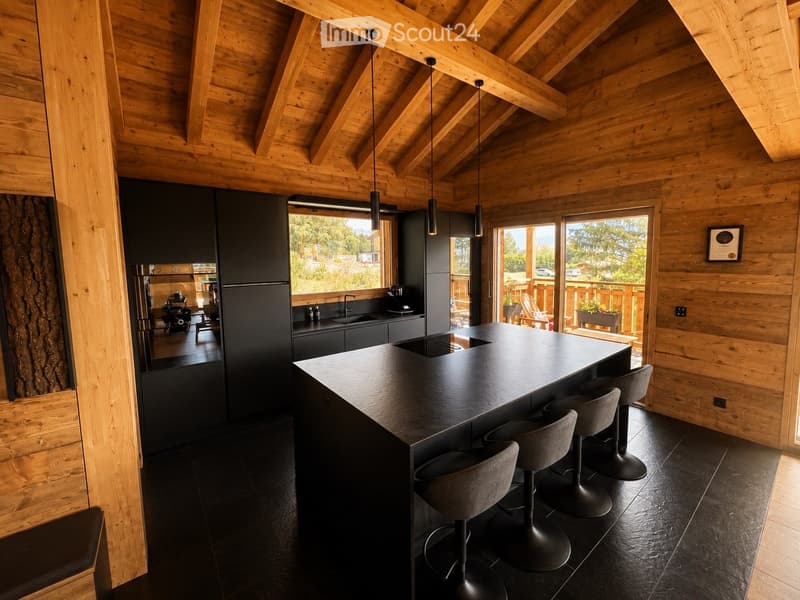 Modern kitchen with wooden beams, black countertops, and a central island with four stools. The space features a sleek refrigerator, sink, and cabinets, with large windows offering a scenic view of the outdoors.