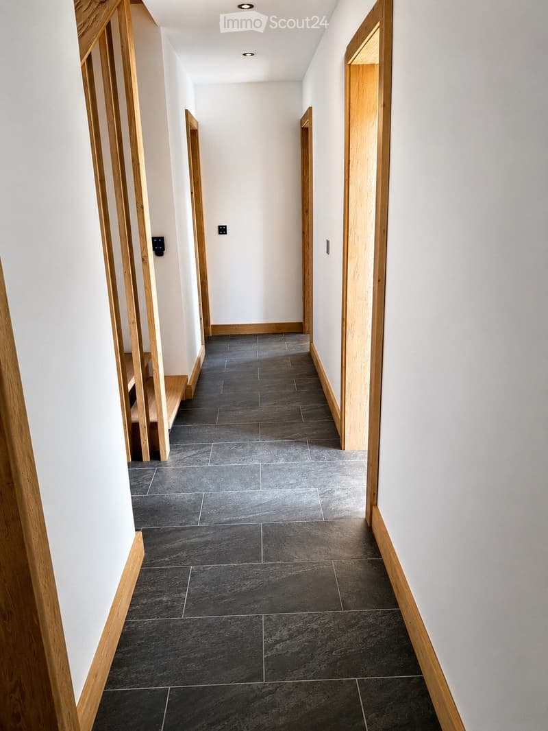 Modern hallway with dark grey tiled floor, white walls, and wooden door frames. Minimalist design with recessed lighting and electrical outlets visible.