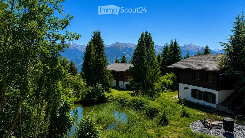 A serene garden area with two houses, surrounded by lush greenery and a small pond, with a backdrop of mountain peaks under a clear blue sky.