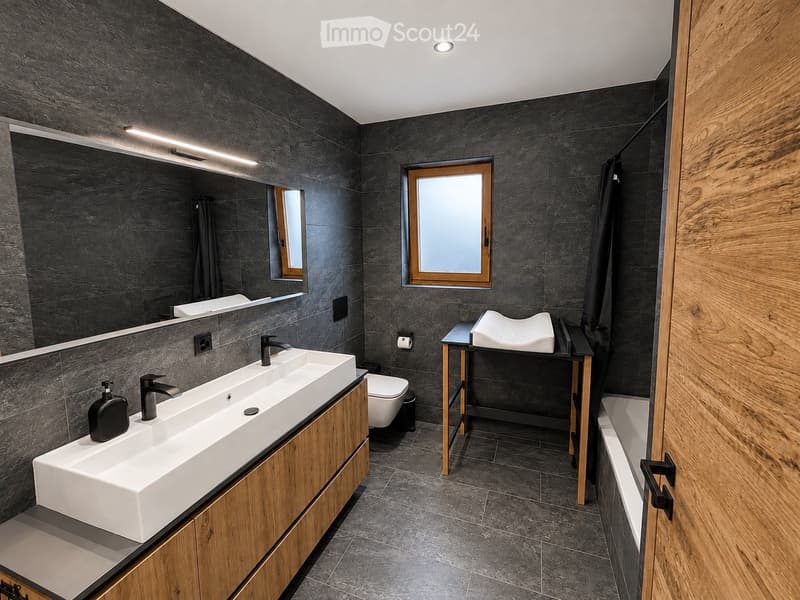 Modern bathroom with dark grey tiles, double sink, large mirror, toilet, small table with stool, and bathtub.