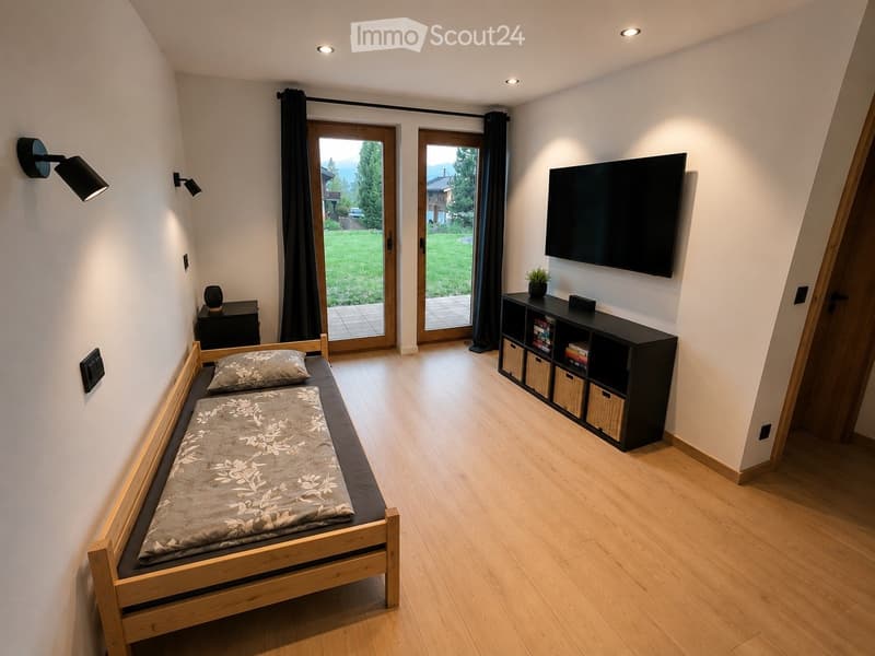 Cozy bedroom with a wooden bed, nightstand, wall-mounted TV, storage unit, and a potted plant. Large windows with black curtains lead to a patio with a view of a green lawn.