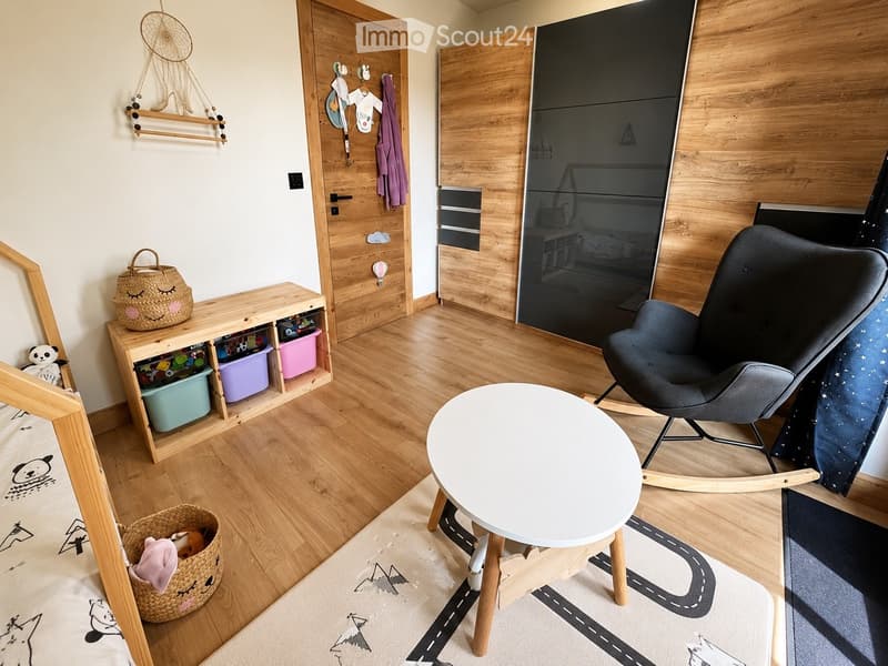A cozy, well-lit children's room with wooden flooring, a round white table, a modern rocking chair, a wooden shelf with colorful storage bins, and decorative baskets.
