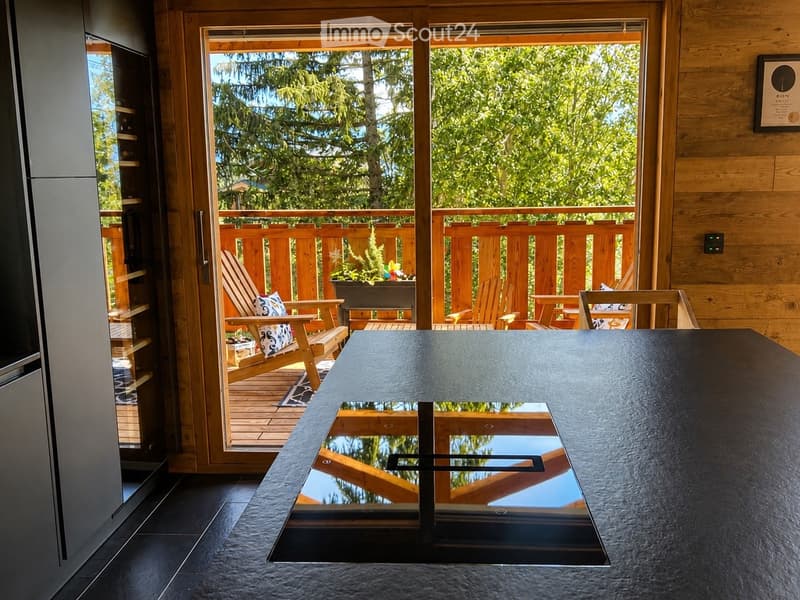 Modern interior with a large glass table and chairs, leading to a wooden balcony with outdoor seating and a view of greenery.
