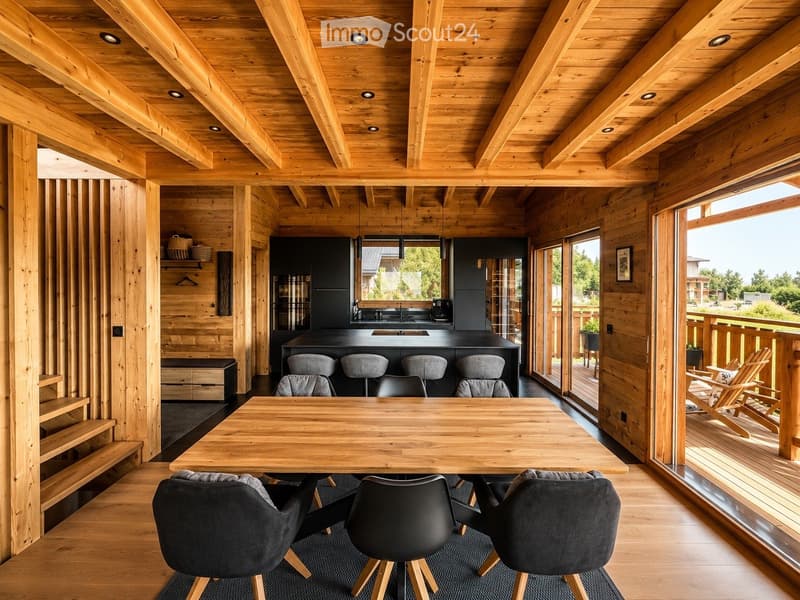 Open-concept living and dining area with exposed wooden beams, a wooden dining table with six chairs, a kitchen island with four stools, and a view to a wooden deck with outdoor seating.