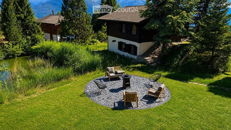 A serene garden area with a circular gravel seating area, featuring lounge chairs and a small table, surrounded by lush green grass and trees, with a pond nearby and a house with a staircase leading up to it in the background.