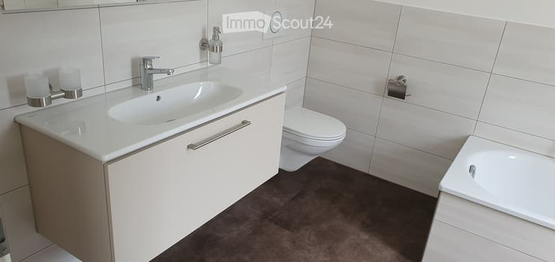 Modern bathroom with a white sink, chrome faucet, toilet, bathtub, and soap dispenser. The walls are tiled in light grey, and the floor is dark brown.