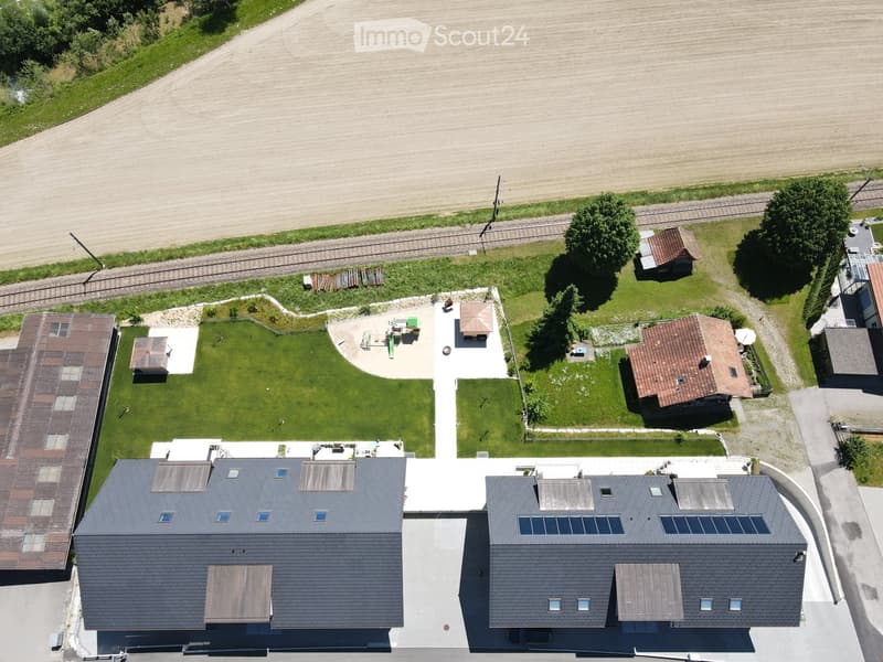Aerial view of a modern house with a grey roof and solar panels, surrounded by a well-maintained lawn, small outbuildings, and a rural landscape with fields and trees.