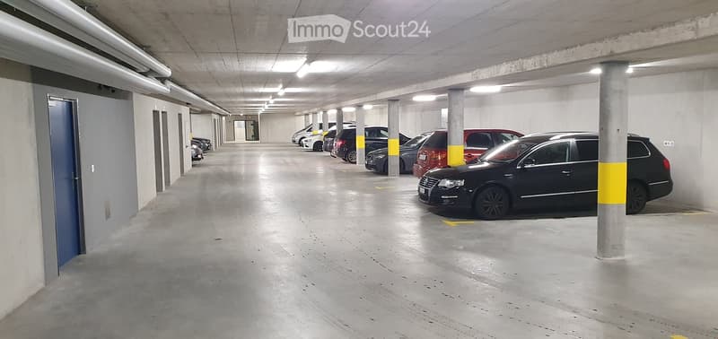 Underground parking area with several parked cars, concrete pillars, and fluorescent lighting.