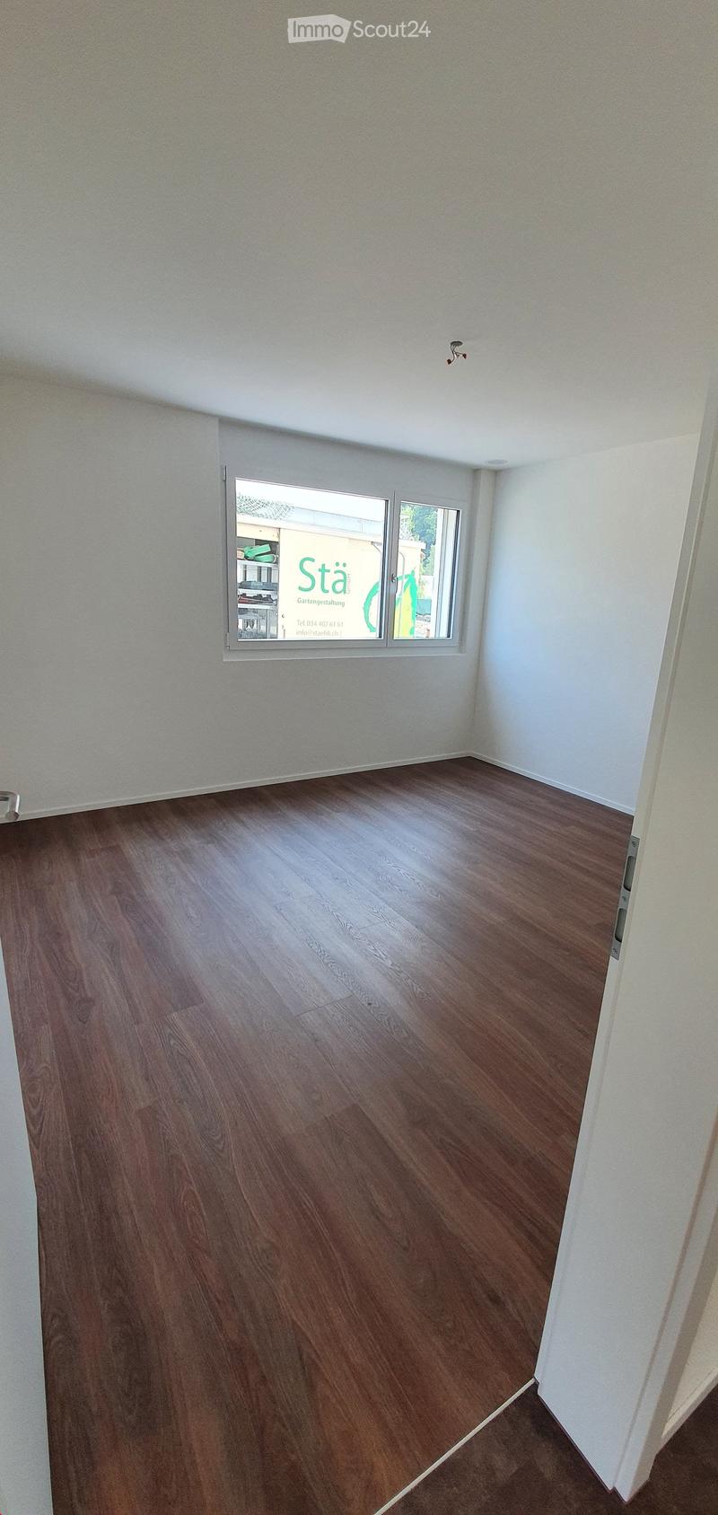 Empty room with wooden flooring and white walls, featuring a large window with a view of a neighboring building.