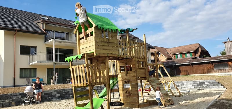 Outdoor play area with a large wooden play structure, including slides, climbing elements, and a swing, surrounded by sand and stone steps. A woman and children are present, with a stroller nearby.