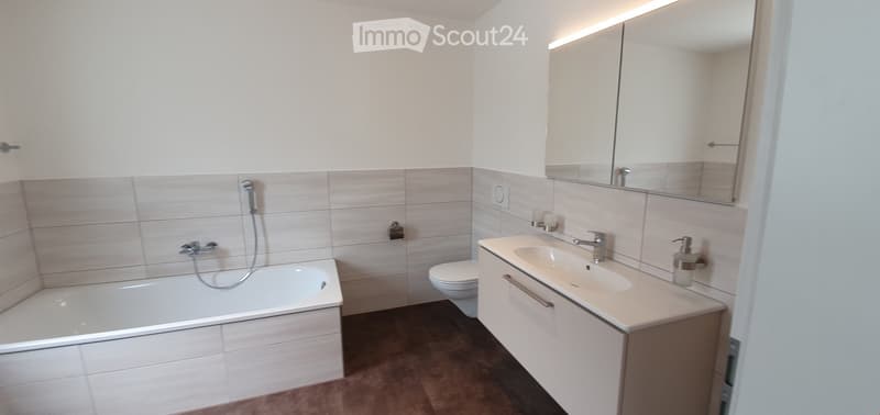 Modern bathroom with a bathtub, toilet, sink, and large mirror. Light-colored tiles and white cabinetry.