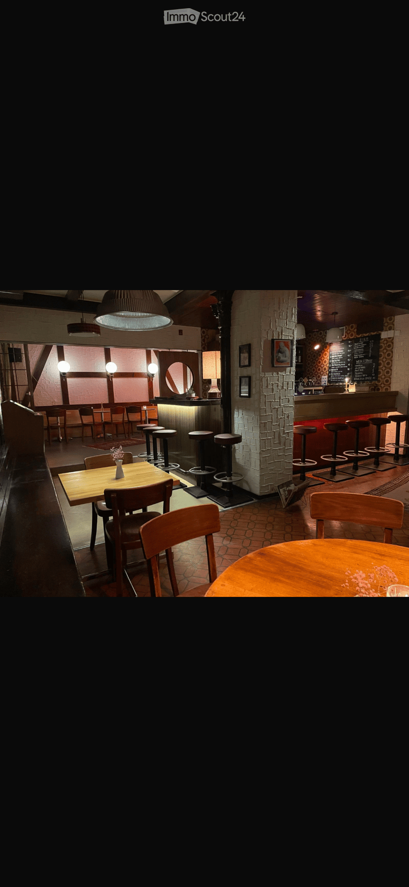 Cozy restaurant interior with wooden tables, chairs, bar stools, pendant lights, and a bar area with a menu board.