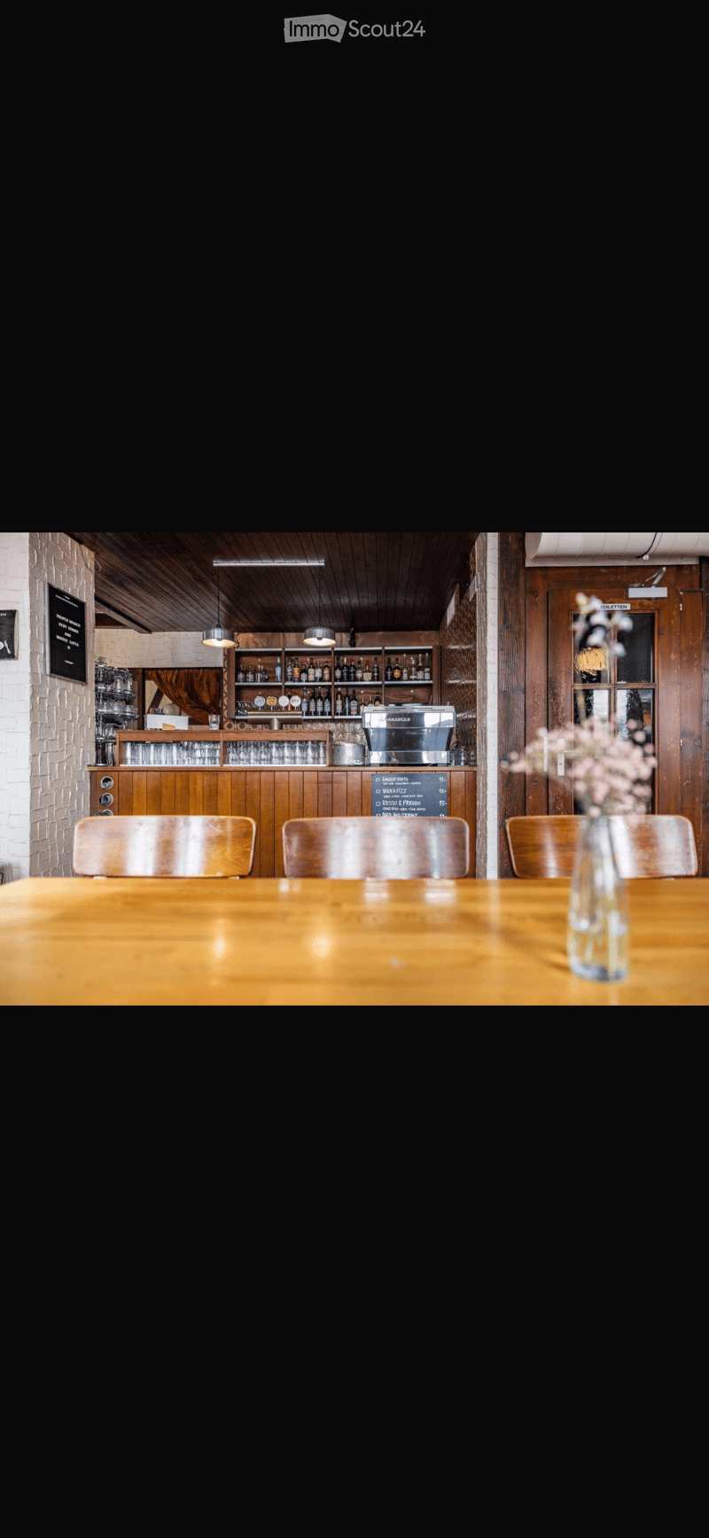 A cozy restaurant interior with wooden tables, chairs, a bar area with shelves of bottles and glasses, and a coffee machine. A vase with flowers is placed on the table.