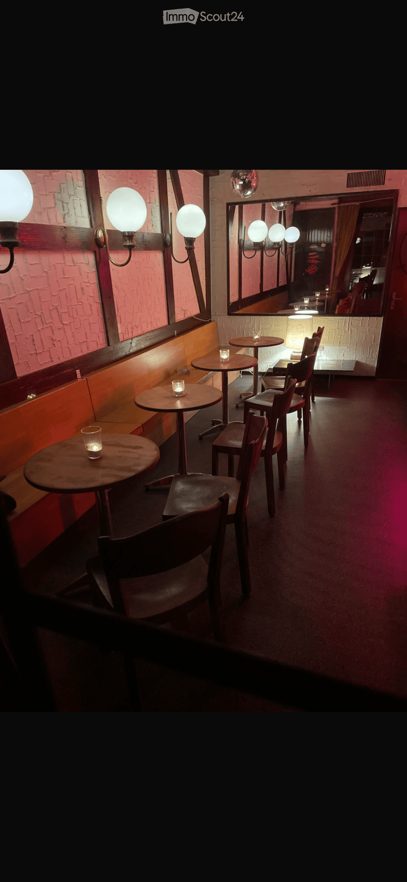 A cozy, dimly lit room with round tables, chairs, a bench seating area, and wall-mounted lamps. The walls are pink brick, and there is a large mirror reflecting the seating area.