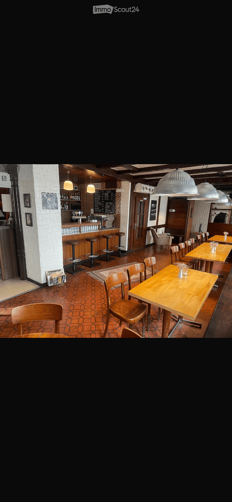 A cozy restaurant interior with wooden tables, chairs, bar stools, and a bar area with a faucet and cups. The space features exposed wooden beams, pendant lights, and a mix of seating arrangements.