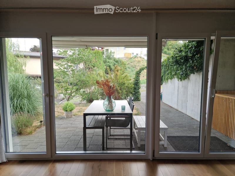 A modern balcony with a glass table, chairs, a vase with dried flowers, and a cup, overlooking a green garden with trees and a pathway.