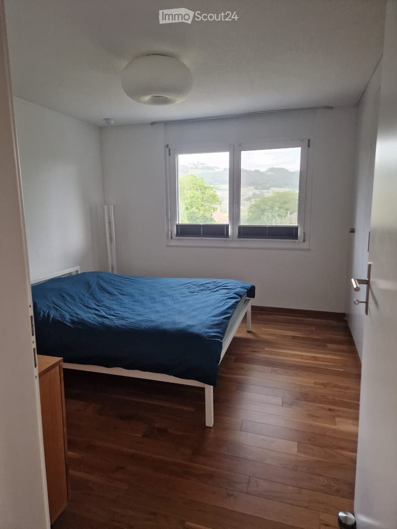Minimalist bedroom with wooden flooring, a single bed with blue bedding, a small nightstand, and large windows offering a scenic view.
