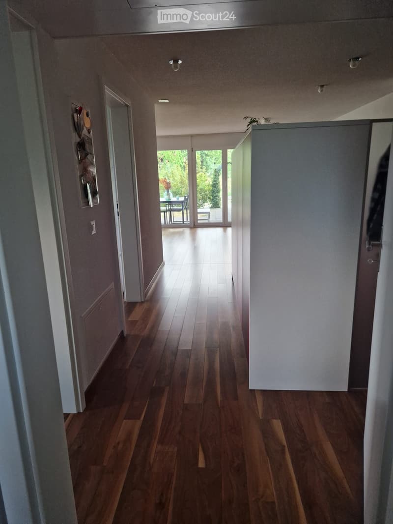 A long, well-lit hallway with wooden flooring, white walls, and several doors leading to different rooms. The hallway features recessed lighting and a small decorative display on the left wall.