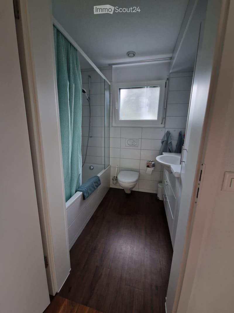 Modern bathroom with a bathtub, toilet, sink, and shower area. Features wooden flooring, tiled walls, and a window with frosted glass.