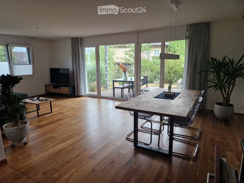 Open-plan living and dining area with wooden flooring, large sliding glass doors leading to a patio, a dining table with chairs, a coffee table, a TV, and potted plants.