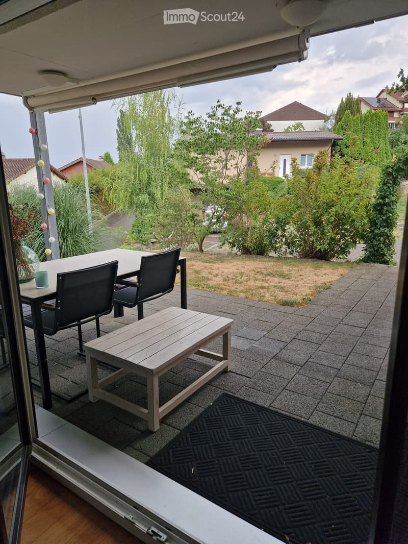 Balcony with a table, two chairs, a wooden bench, and a cup, overlooking a garden with trees and houses.