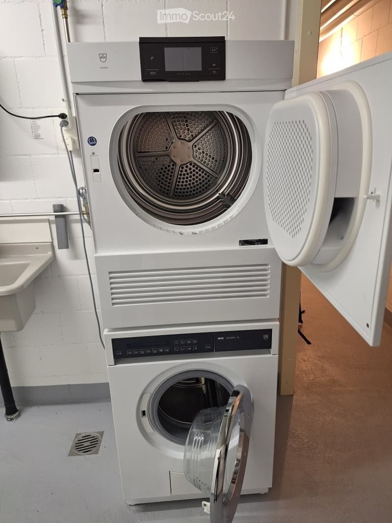 A compact laundry area with a stacked washing machine and dryer unit, both white with control panels, placed against a tiled wall with a small sink to the left.