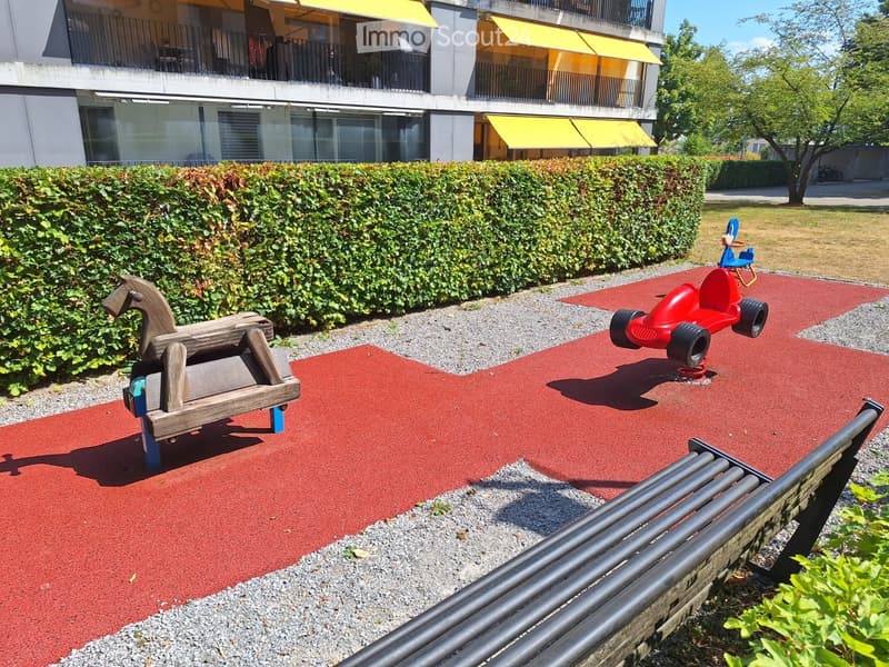 Playground area with a wooden rocking horse, red toy car, and a bench, surrounded by a hedge and a modern building with balconies.