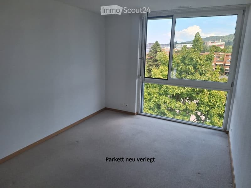 Empty room with light-colored walls, carpeted floor, and large window offering a view of trees and buildings.