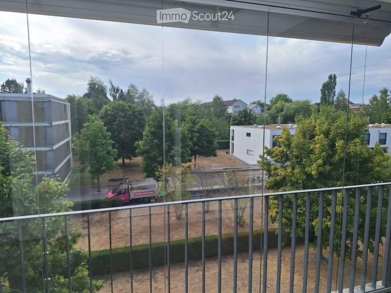 Balcony with a metal railing, overlooking a landscaped area with trees, buildings, and a parked van.