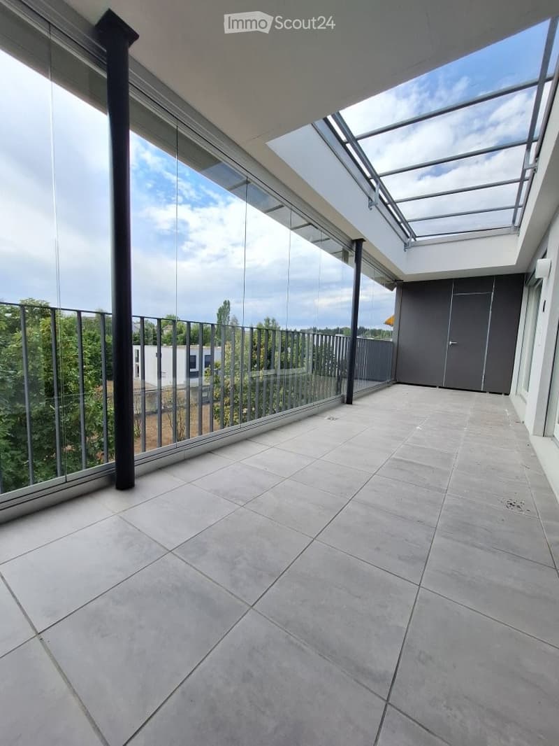 Modern balcony with large glass panels, tiled floor, and a view of greenery outside.
