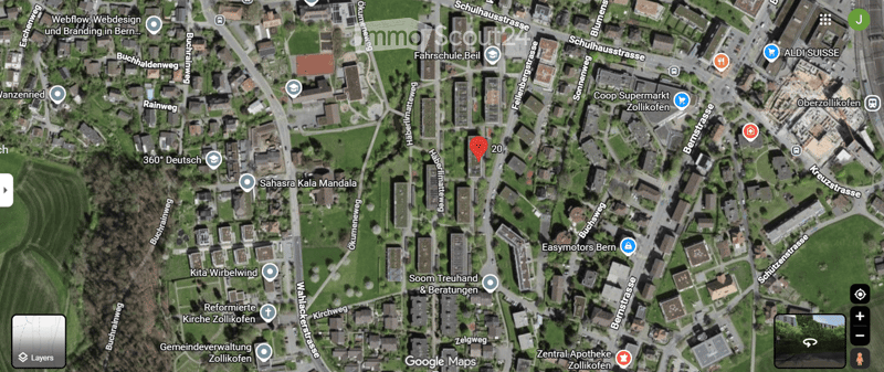 Aerial view of a residential area with multiple buildings, streets, and green spaces. Marked location at the center indicates a specific point of interest.