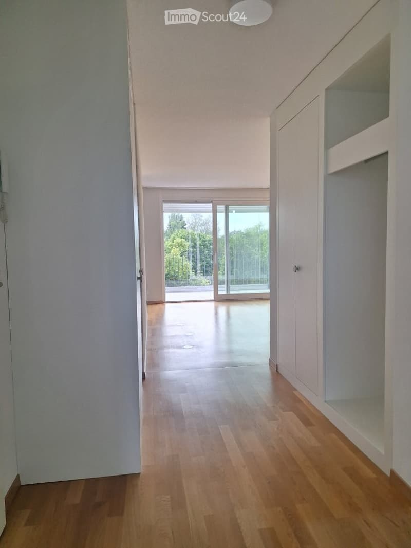 Empty hallway with wooden flooring, white walls, and large glass doors leading to an outdoor area.