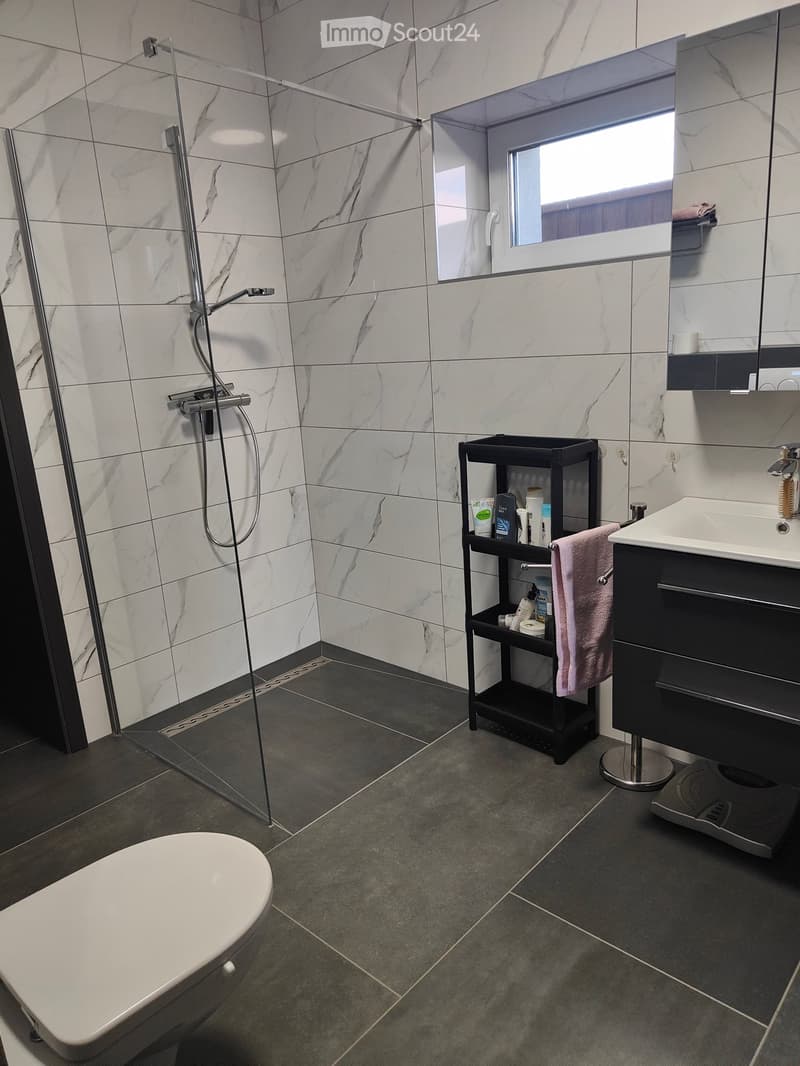 Modern bathroom with marble tiles, a glass-enclosed shower, a white toilet, a black vanity with a sink, a mirror, and a small black shelf holding toiletries and a pink towel.