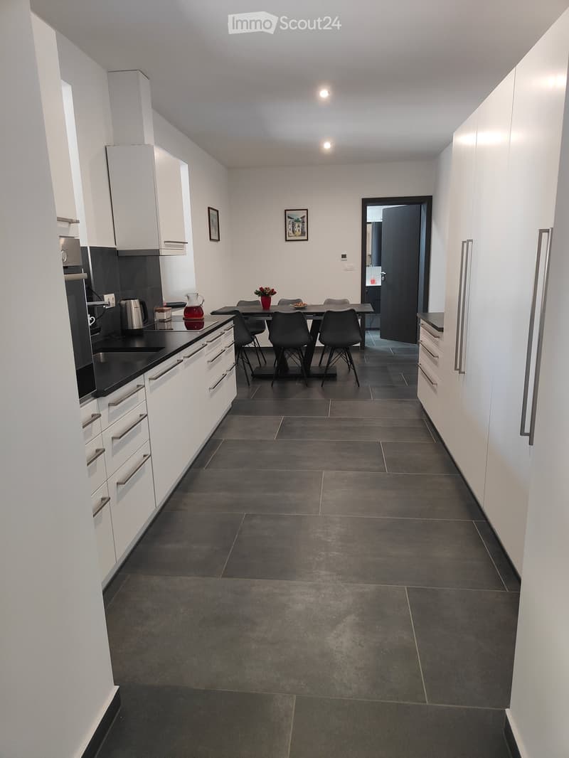 Modern kitchen with white cabinets, dark countertops, and a dining area with a table and chairs. The floor is tiled, and there is a vase with flowers on the table.