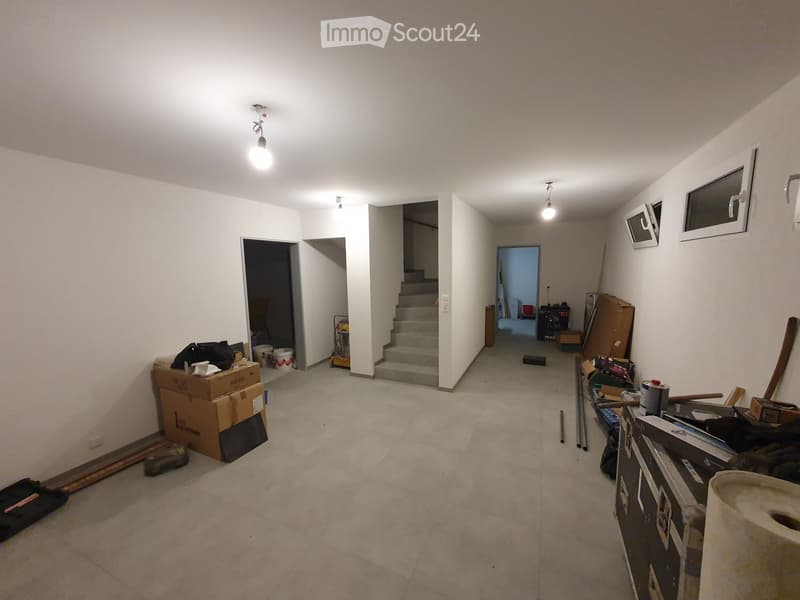 Empty hallway with white walls, concrete floor, and a staircase leading upwards. Various construction materials and tools are scattered around, including boxes, a ladder, and a workbench.