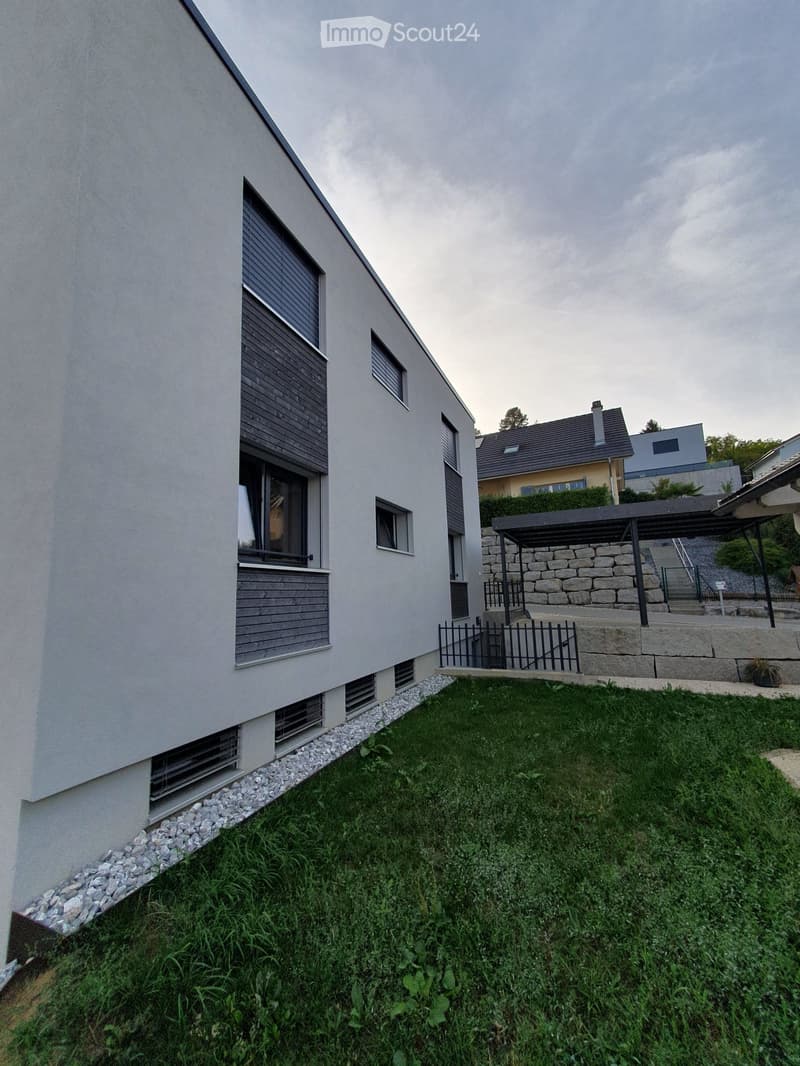Modern two-story house with grey exterior, multiple windows, and a small garden area with gravel border and lawn.