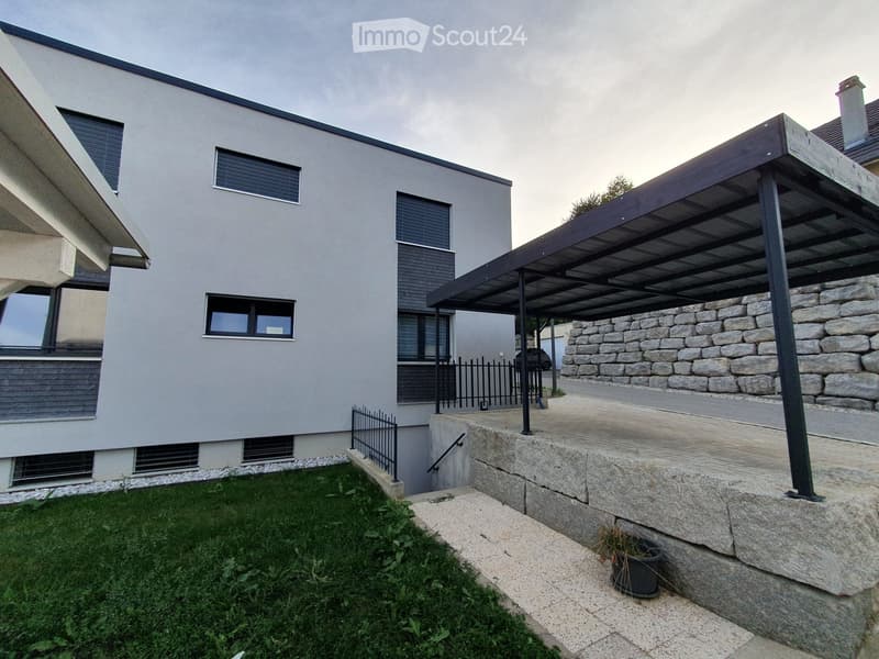 Modern two-story house with grey facade, large windows, raised ground floor, stone retaining wall, covered parking area, and small garden.