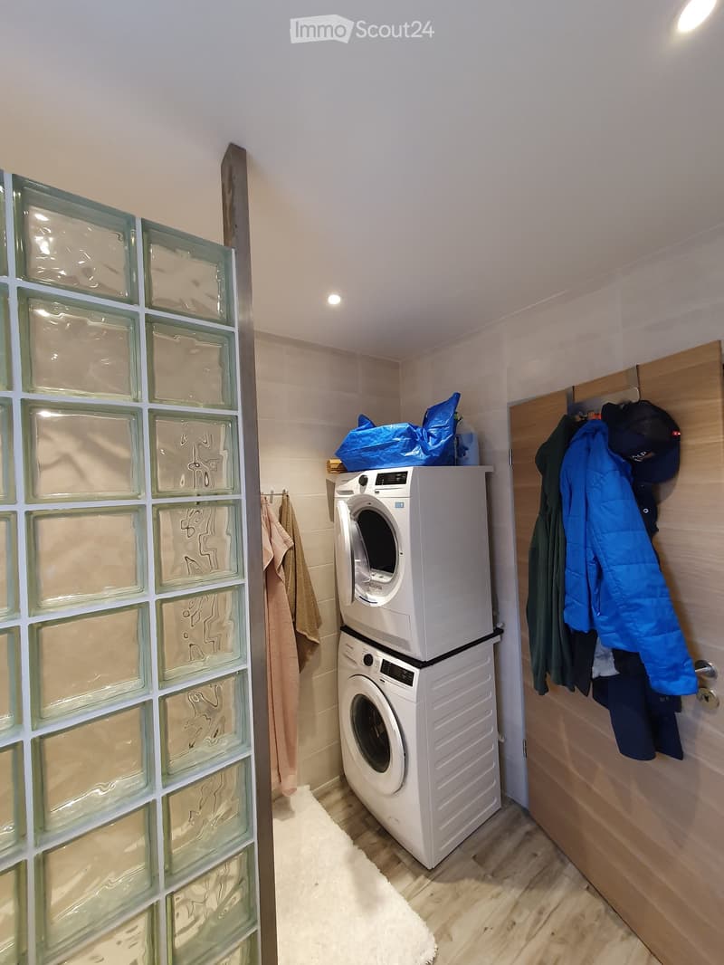Compact bathroom with a stacked washing machine and dishwasher, wooden floor, glass block wall, and hanging coats and towels.