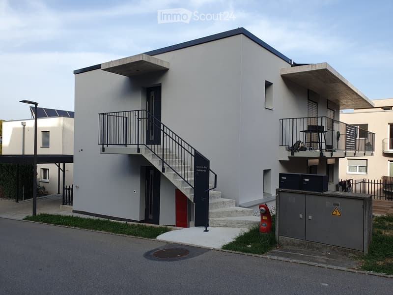 Modern two-story building with grey facade, black railings, and a staircase leading to the upper level. Features include a small balcony, a red utility door, and a utility box with a warning sign.
