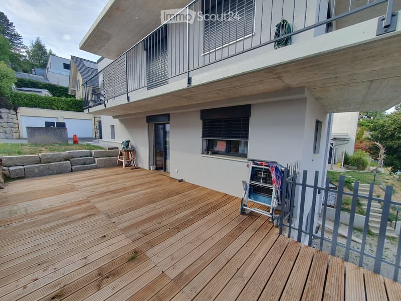 Wooden deck with modern railing, adjacent to a house with large windows and a balcony above.
