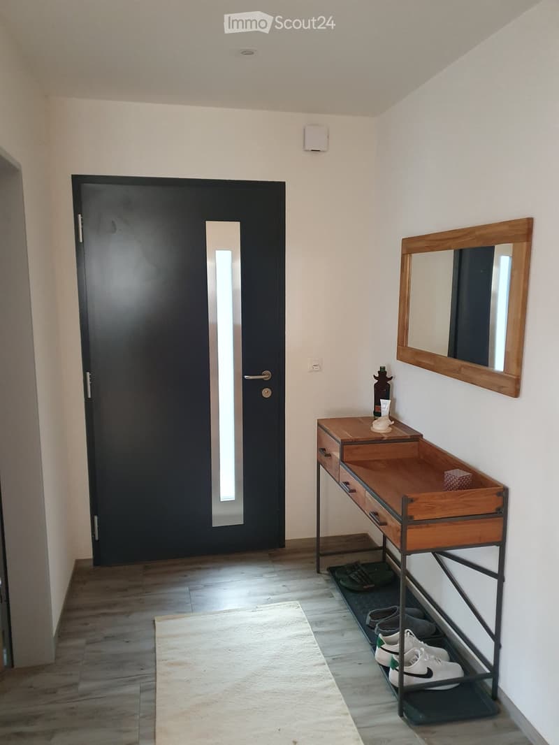 A modern hallway with a wooden floor, a black door with a glass panel, a small wooden console table with a mirror, and a shoe rack holding several pairs of shoes.
