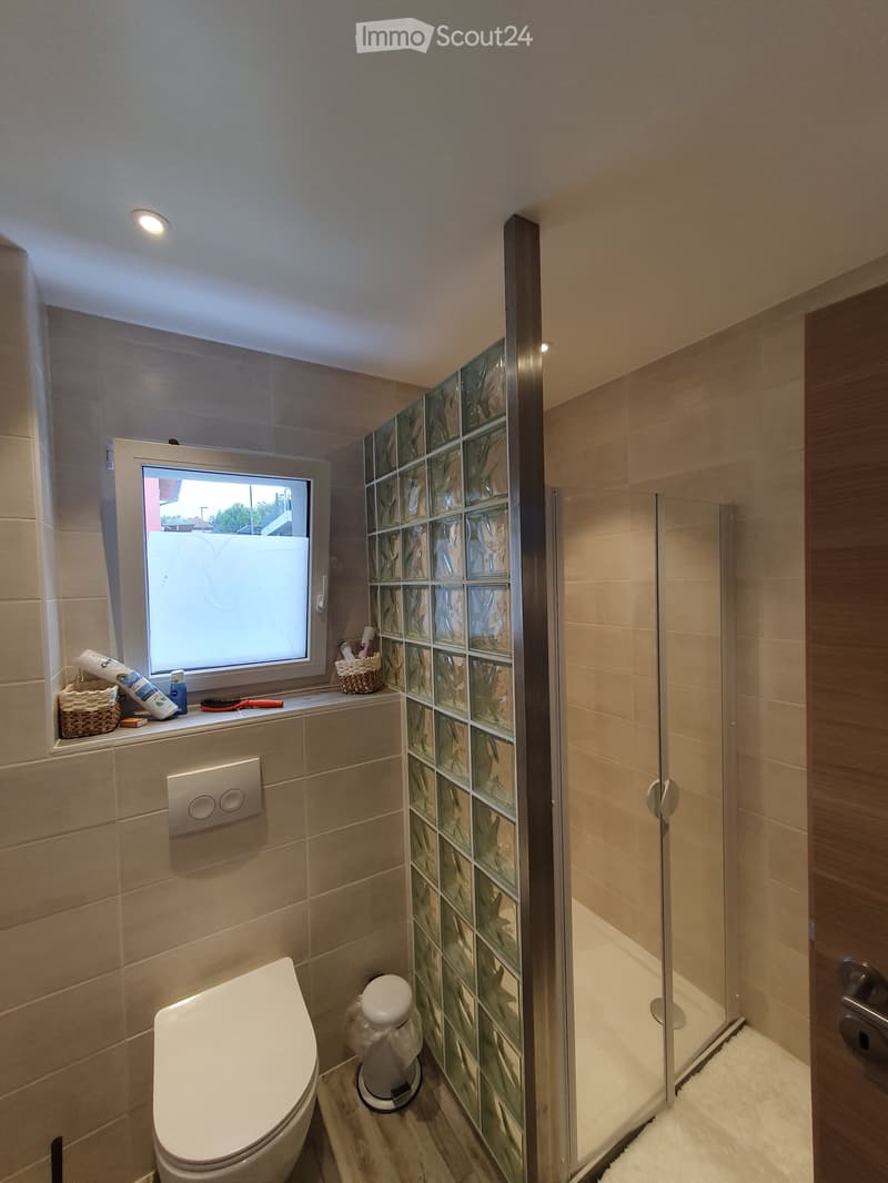 Modern bathroom with beige tiles, a toilet, a glass-enclosed shower, a window with frosted glass, and a small wicker basket on the windowsill.