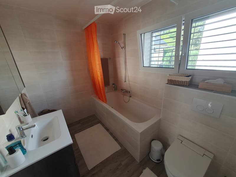 Modern bathroom with a white sink, bathtub, toilet, and mirror. Features a window with blinds, a shower hose, and a small basket on the windowsill.