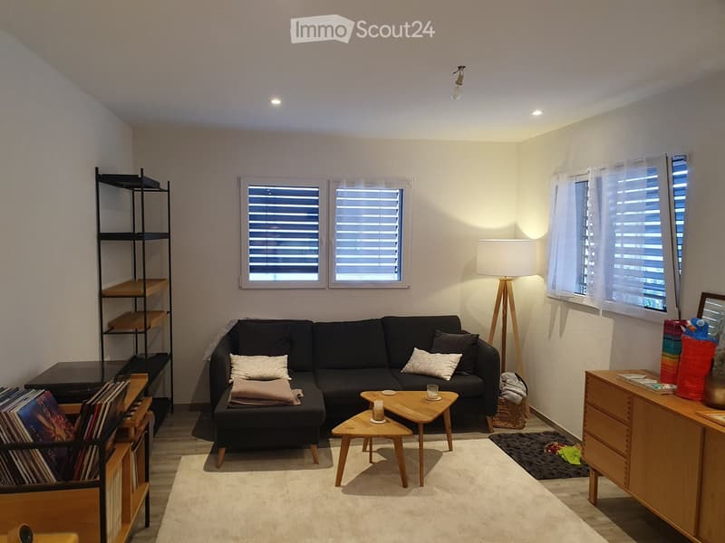 Modern living room with a black sectional sofa, wooden coffee table, floor lamp, and shelves. The room features large windows with blinds and a cozy carpeted floor.