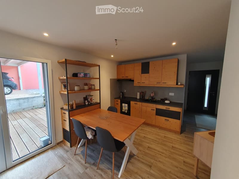 Modern kitchen with wooden cabinets, a central dining table with chairs, and a large open shelving unit. The space features a sleek, contemporary design with integrated appliances and ample natural light from the large window.