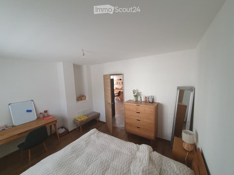 Cozy bedroom with a bed, desk, chair, mirror, lamp, and a small bench. The room features wooden flooring and white walls, with a partially visible adjacent room through an open door.