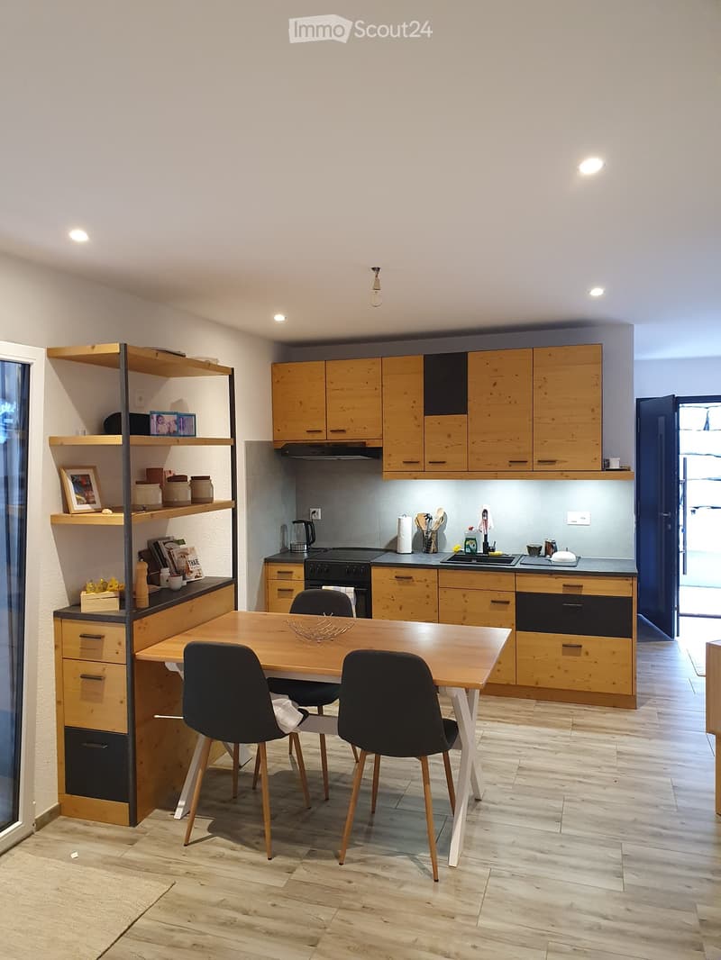 Modern kitchen with wooden cabinets, a central dining table with four chairs, and various appliances including an oven, sink, and countertop items.